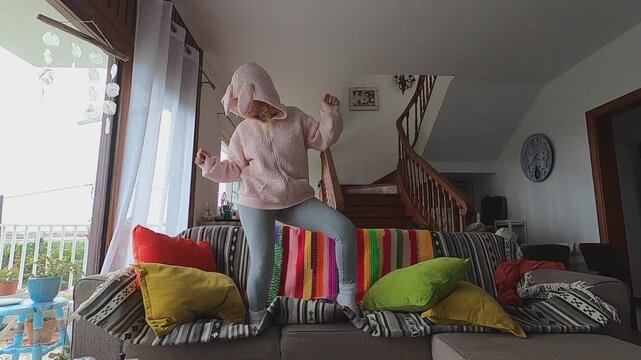 A joyful young girl in a cozy pink bunny ear hoodie enthusiastically dances and jumps with playful energy on the living room sofa, radiating carefree happiness and fun at home.