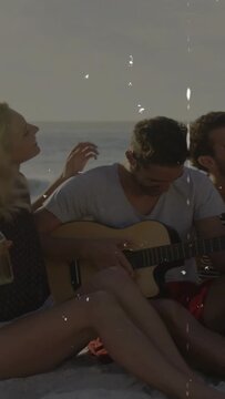 Vertical video: Guitarist starting strumming on beach, sparking friends laughing with bottle raised