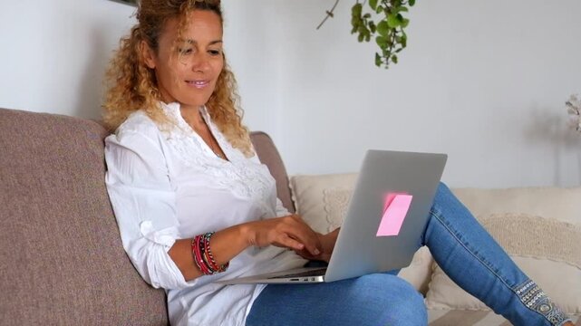 A content woman with curly hair enjoys a productive remote work session from the comfort of her home, smiling while typing on her laptop on the cozy couch, embodying modern flexible work.
