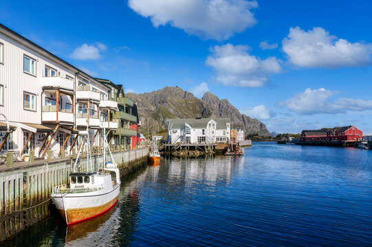 Picturesque harbor view of Henningsv&aelig;r on the Lofoten Islands with fishing boat, traditional wooden houses, calm blue water and impressive rocky mountains under sunny autumn sky.