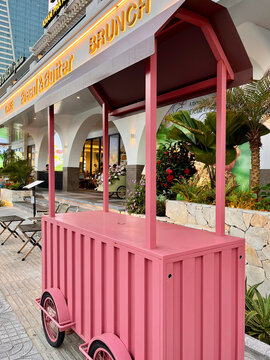 A vibrant pink food cart is positioned outdoors, featuring a canopy and two rounded wheels. Surrounding greenery adds to the lively atmosphere. Ideal for brunch scenes.