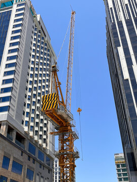 A tall construction crane stands between two modern skyscrapers in an urban environment. The crane is yellow with a striped warning pattern. It is clear and sunny.