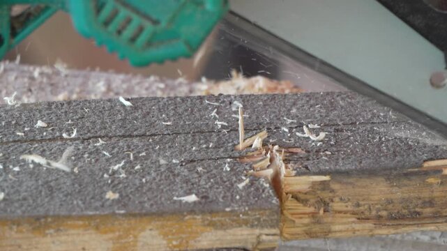 Electric chainsaw blade slices deep notch into rough wooden board, scattering sawdust across workshop floor. High angle close up shot.