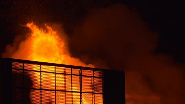Bright orange flames surge upward behind black metal mesh as dense smoke rolls through a dark interior, illuminating overhead structures with intense flickering firelight