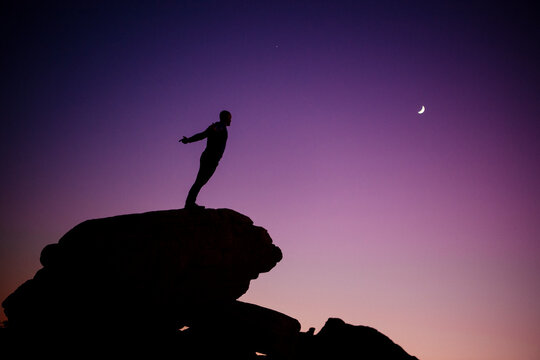 Silhouette of white man standing on rock under crescent moon