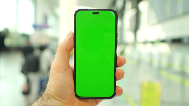 Hand holds phone with green screen in airport. Background shows blurred travelers and terminals. Phone screen is solid green for overlay. Hand visible with natural skin tone