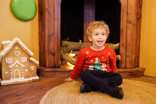 Little boy wearing a Christmas sweater sitting by a fireplace, smiling and feeling the festive cheer