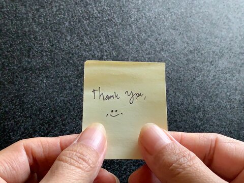 A person holding a yellow sticky note with handwritten Thank You text and a smiley face using both hands over a dark textured background in Jakarta, Indonesia