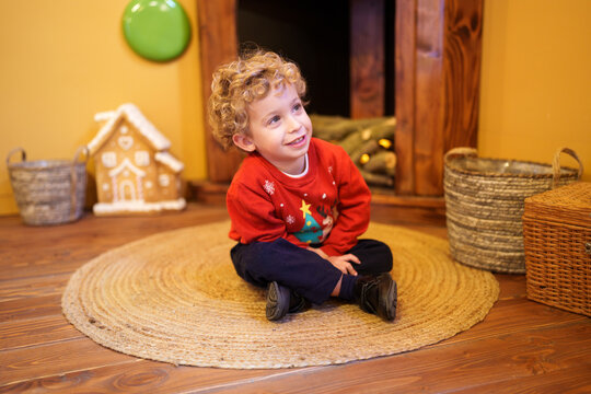 Young boy smiling, sitting on rug, wearing festive Christmas sweater, looking up with joy during holiday season