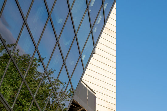 Reflective glass facade with tree reflections beside a white wall, showing modern architecture through geometric glass and reflections across the facade