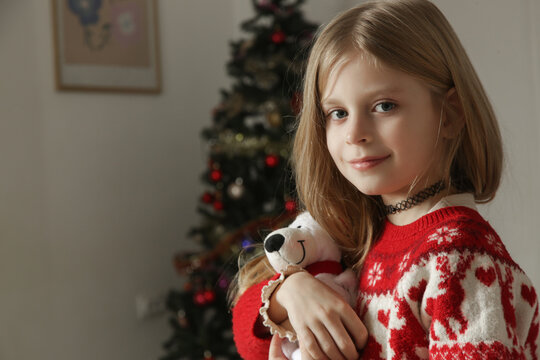 Side view profile of a cute blonde child sitting on a rug and gently holding a plush teddy bear with a Santa hat. Young girl wearing a red festive winter jumper with reindeers, cozy holiday atmosphere