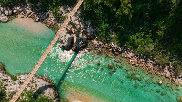 Aerial top down perspective captures turquoise Soca river crystal clear water. Simple wooden footbridge crosses rocky canyon banks surrounded by dense wild green forest near Kobarid.