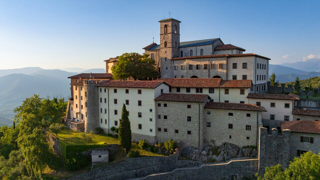 Majestic drone shot displays historic sanctuary of Castelmonte on hilltop. Ancient stone fortification walls and religious architecture stand over Friuli region landscape under clear sky.