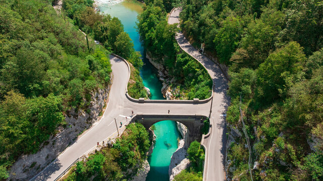 Aerial top down perspective captures turquoise Soca river crystal clear water. Simple bridge crosses rocky canyon banks surrounded by dense wild green forest near Kobarid.