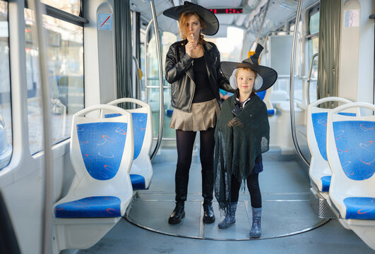 Mother and daughter wearing Halloween witch hats board city tram after seasonal park walk. Family holds jack o lantern pumpkin on autumn city street.