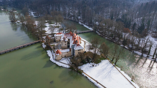 Picturesque drone panorama shows historic Otocec Castle covered in light snow. Frozen park trees, wooden bridges, and cold green Krka river frame medieval island palace in winter time.