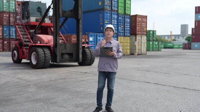 Wide shot of a warehouse officer noting cargo details on a clipboard in an open container yard filled with colourful boxes and a stacker in a busy container depot.