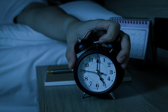 Hand turning off alarm clock on bedside table in dark bedroom. Concept of early morning wake up, insomnia, time management, sleep disorder, and routine.