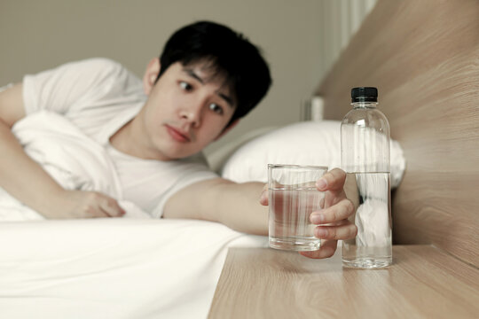 Asian man lying on bed reaching for glass of water on table. Concept of morning routine, hydration, healthy lifestyle, thirst, and waking up.