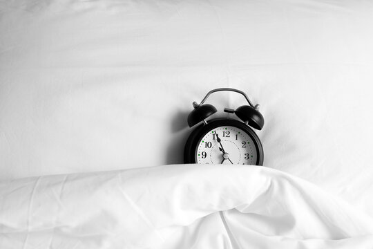 A black alarm clock resting under a white duvet on the bed. Time management concept.