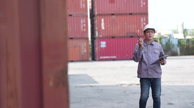 A terminal coordinator calls on a two way radio while holding a tablet, then walks past the camera between tall container rows at a busy container terminal yard.