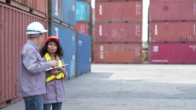 Two logistics staff walk slowly along a container wall while comparing figures on a tablet and talking through the loading plan at a busy container terminal yard.