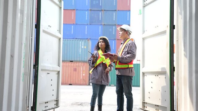 A female inspector and her supervisor stand between the open steel doors of a shipping container checking a tablet and a radio at a busy container terminal yard.