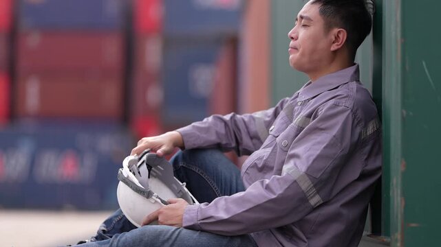 An exhausted container yard worker sits on the ground against a container with his helmet off, rubbing his eyes after a long shift at a busy container terminal yard.
