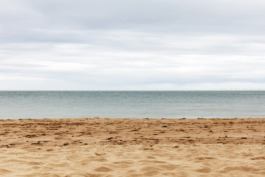 Quiet sandy beach beside a calm sea under a soft gray cloudy sky, with a wide open horizon and empty shoreline.