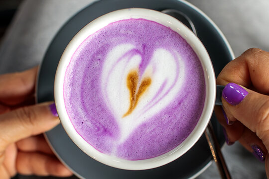 Hand with purple manicure holding dark ceramic cup of purple ube latte topped with heart shaped latte art foam, cozy coffee shop atmosphere closeup photography shot today, high quality photo