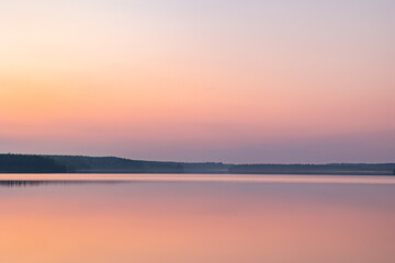 Obraz premium Peaceful sunset reflections over a calm Finnish lake during twilight in Finland. Soft pastel shades of pink, orange, and purple color the gradient sky and smooth water