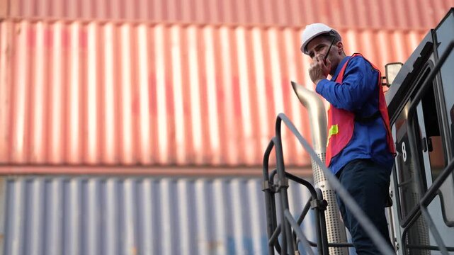 A container handler operator climbs the steel steps to his cab while calling the next lift through on a handheld two way radio at a busy container terminal yard.