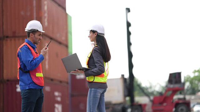 Two logistics professionals walk along the container row with a laptop while a reach stacker lifts a box in the background during a routine shift at the container terminal.
