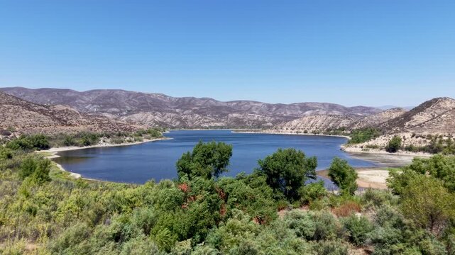 Vail Lake framed by dense shrubs and trees against dry rugged hills beneath a clear blue sky, aerial establishing dolly flyover