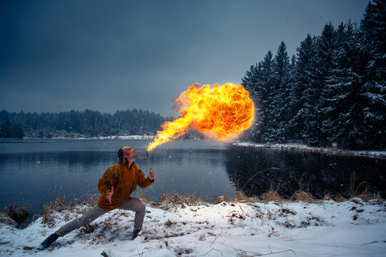 Fire breather spitting a huge fireball outdoors on a snowy winter lake shore with falling snow