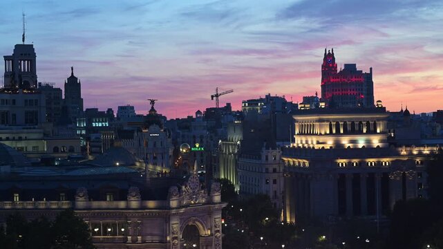Elevated dusk perspective over illuminated Madrid skyline with Bank of Spain and Metropolis building as beautiful sunset pink and red colors glow behind city roofs in Spain, architectural b-roll.