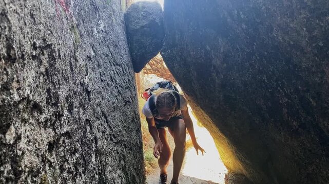 A woman with a backpack bends forward to pass through a narrow gap between large boulders while hiking on Mount Mulanje, Malawi.