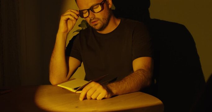 Sad and stressed man sitting at a table, covering his face with his hand while writing in a notebook under dramatic cinematic lighting.