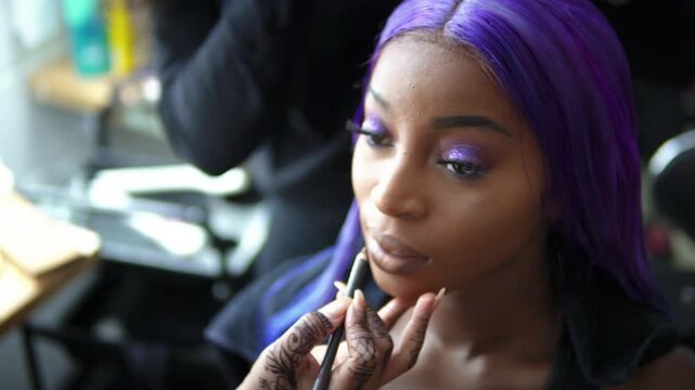 Tight shot focusing on the delicate application of lip liner. The artist's henna-patterned hand gently touches the chin of a gorgeous Black woman sporting a vivid purple wig and heavy glam makeup.