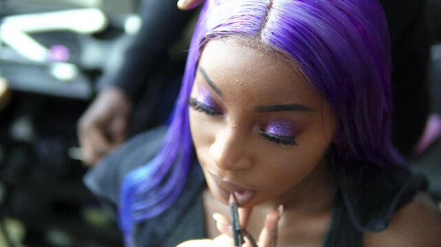 Close-up of a professional makeup artist tattoos applying nude lip liner to a stunning Black model featuring vibrant purple hair and glittery eyeshadow during a studio prep. Brushing her hair slowmo