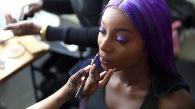 Beauty and cosmetics industry concept. A gorgeous model with striking violet hair and sparkling eye makeup has her lips outlined. The makeup artist's hands intricate traditional henna art. Slow motion