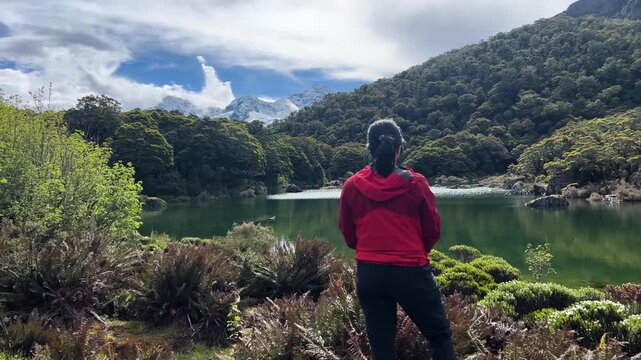 A walker in a red jacket stands at the edge of Lake Mackenzie on the Routeburn Track, New Zealand, green water and beech forest running to a snow capped peak beyond. Wide shot