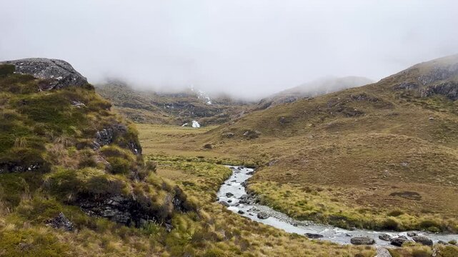 Cloud hides the tops above a tussock basin on the Routeburn Track, New Zealand, a braided stream winding between rocks and golden grass on the valley floor. Pan right shot