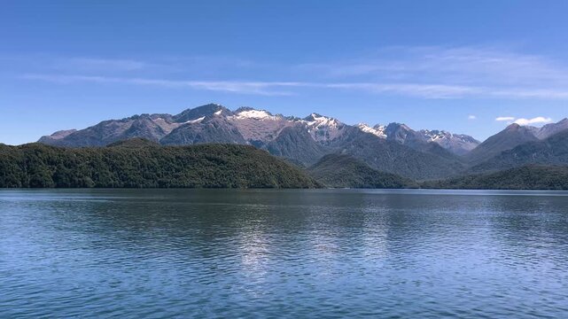 A bush covered shoreline runs along Lake Manapouri in Fiordland, New Zealand, a long snow patched range standing behind it under a clear summer sky. Wide shot