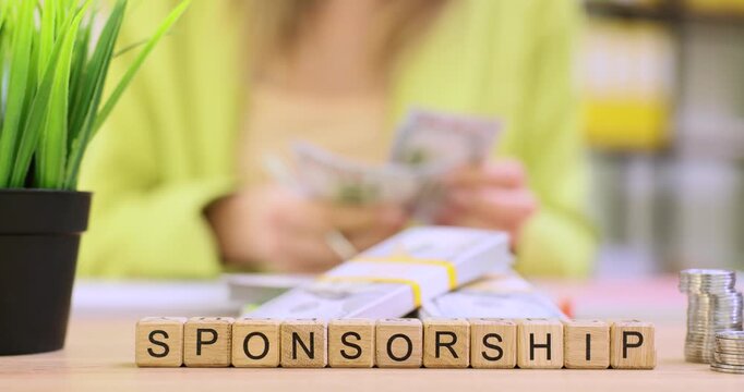 Wooden blocks spell Sponsorship near stacked banknotes wad on desk. Office worker counts cash handling payment bundle carefully in background