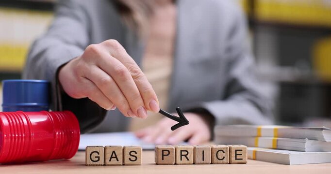 Wooden blocks spell Gas Price near cash stacks and toy barrels on desk. Office worker places black arrow above term highlighting rising fuel costs