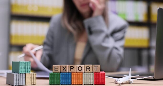 Wooden blocks spell Export put on cargo containers and toy plane on desk. Female office worker talks on phone stamping papers behind trade display