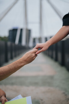 Two people hold hands on a pedestrian bridge, one hand offering support. Close-up of their joined hands conveys connection and intimacy in an urban setting.
