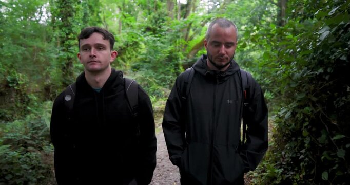 Two men walking through lush forest trail, wearing jackets and backpacks