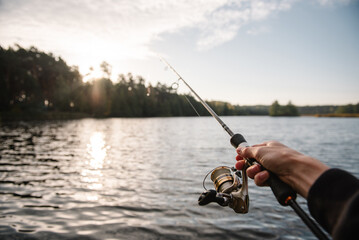 Fototapeta premium First person view of a fisherman holding a spinning rod with a reel over a serene lake at sunset, celebrating Fisherman's Day in nature.